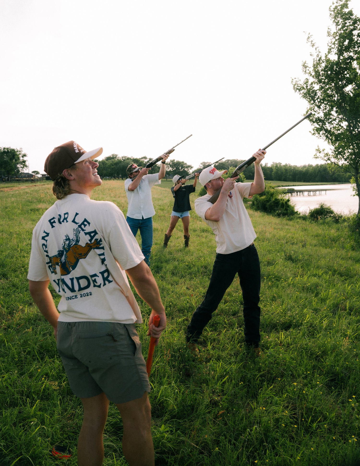 People in a field with one holding a rifle, another with a bow, and a lake in the background.
