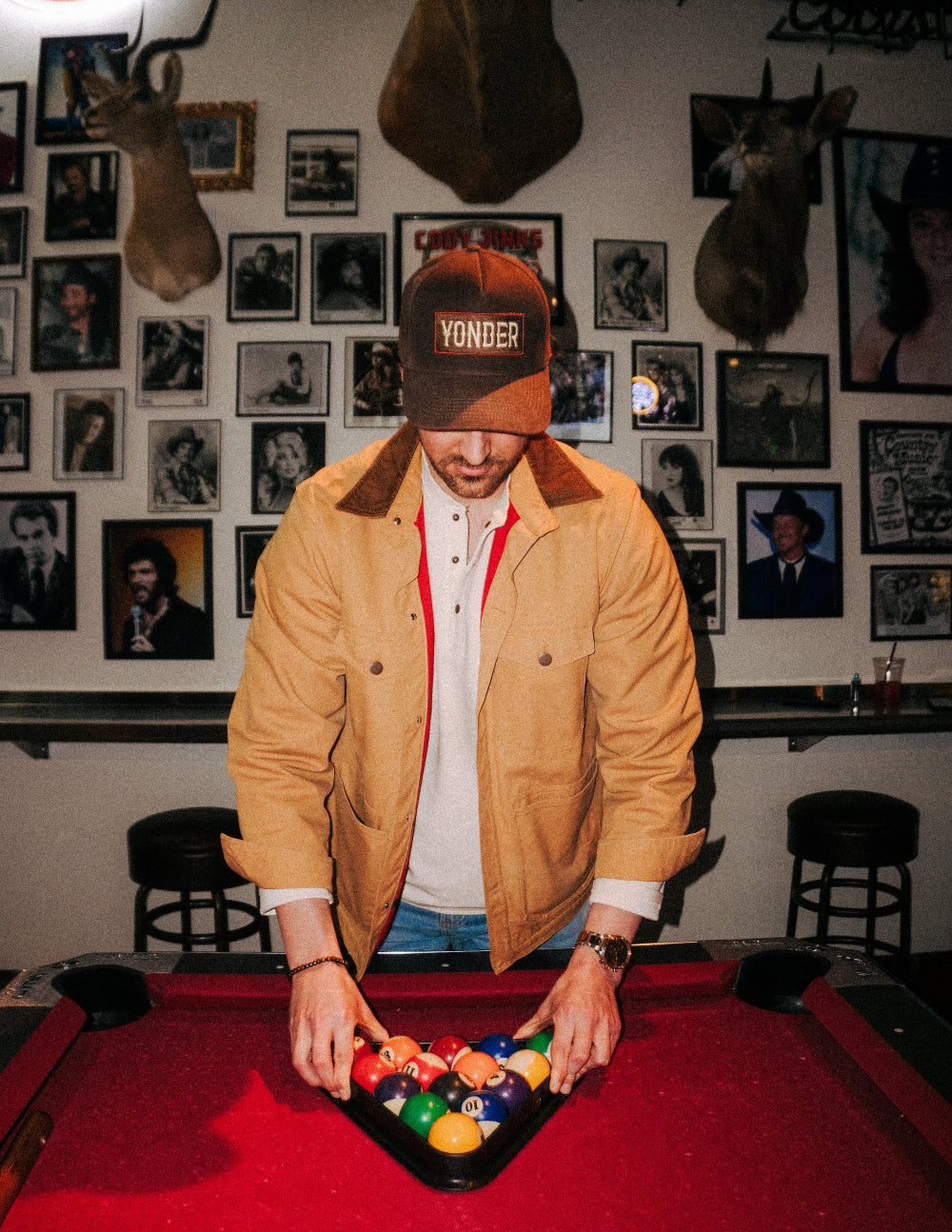 Man in a tan jacket and red cap arranging pool balls on a pool table with a wall of framed photos in the background.